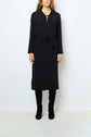 A person wearing a robe longue noire à manches longues with lace-up front, front pockets, a robe ceinturée at the waist, and black knee-high boots, standing against a plain light background.
