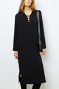 Woman wearing a black, long-sleeve, lace-up robe longue noire with front pockets and a black shoulder bag. The dress features side slits, and she has long, loose hair.