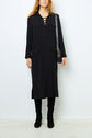 A woman stands against a white wall wearing a robe longue noire à lacets, paired with black knee-high boots and carrying a black shoulder bag. Her hair is loose, and her hands are relaxed by her sides.
