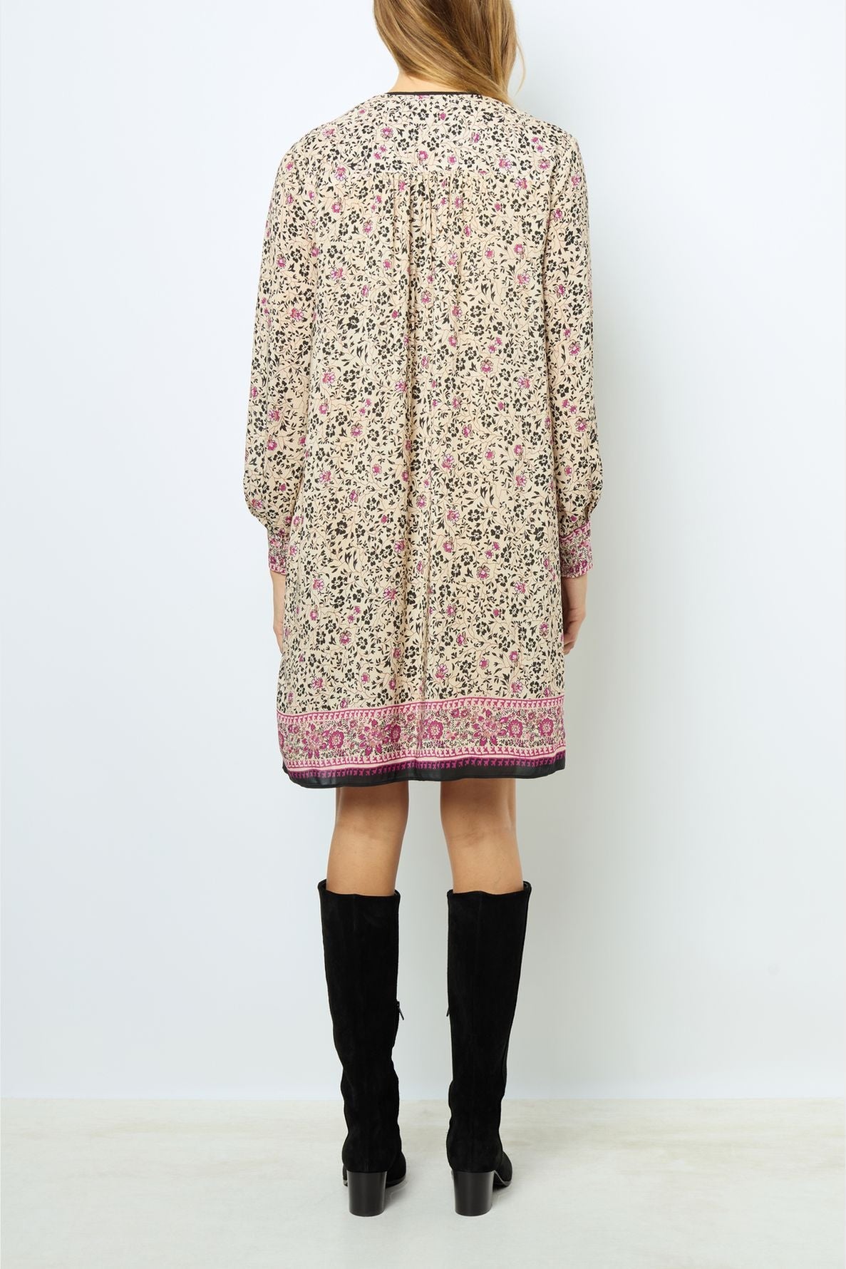 A woman stands facing away, wearing a robe courte à manches longues, cream-colored with an imprimé floral and a pink border, paired with knee-high black boots.