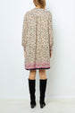A woman stands facing away, wearing a robe courte à manches longues, cream-colored with an imprimé floral and a pink border, paired with knee-high black boots.