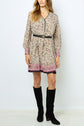 A woman wears a long-sleeved, patterned beige dress with a black belt and pink accents, featuring un élégant imprimé floral, paired with tall black boots, standing against a plain white background.