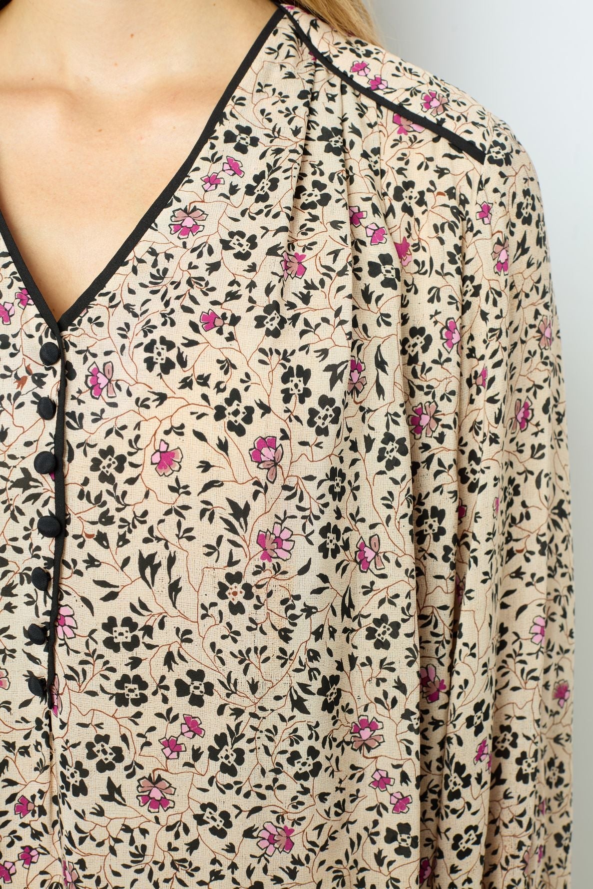 Close-up of a beige blouse with an imprimé floral in black and pink, featuring black trim along the collar and neckline, and black buttons. Part of a person’s neck, shoulder, and chest are visible.