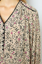 Close-up of a beige blouse with an imprimé floral in black and pink, featuring black trim along the collar and neckline, and black buttons. Part of a person’s neck, shoulder, and chest are visible.