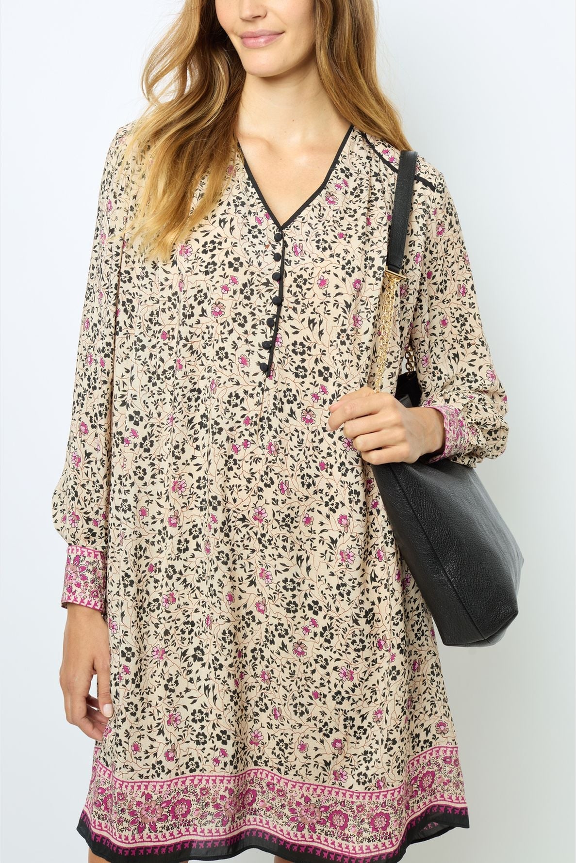 Woman wearing a robe courte à imprimé floral with pink and black accents and long sleeves, holding a black shoulder bag. She has long, light brown hair and is standing against a plain light background.