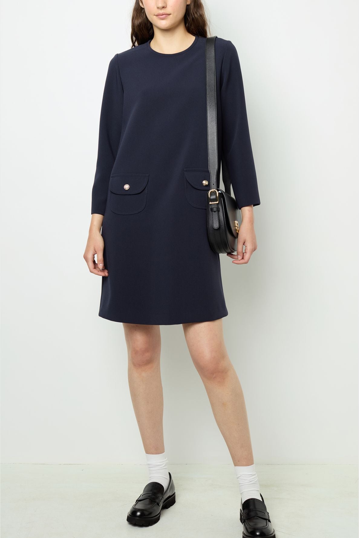 Woman wearing a navy long-sleeve robe courte with deux poches frontales et des boutons dorés, paired with a black shoulder bag, white socks, and black loafers, standing against a plain white background.