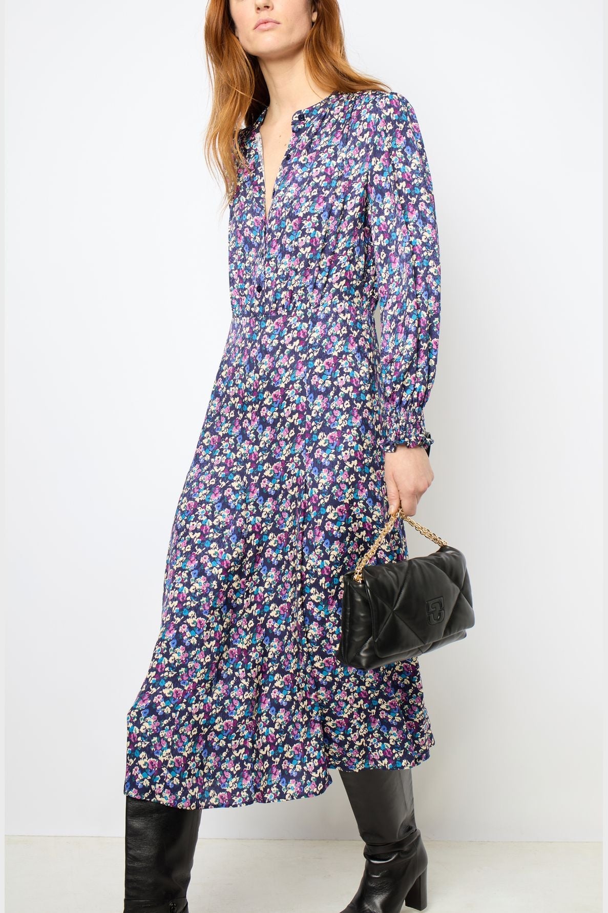 Woman in a long-sleeve, blue jacquard robe midi dress with a V-neck and motif floral design, holding a black quilted handbag with a chain strap, and wearing black knee-high boots. The background is plain white.