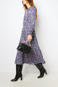 Woman in a long, blue motif floral robe midi and black knee-high boots holds a black handbag and walks against a plain white background.