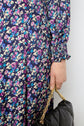 A person in a long-sleeve, purple motif floral jacquard dress holds a black quilted handbag with a gold chain strap. Only the lower arm and hand are visible against a neutral background.