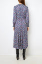 A person stands facing away, wearing a long-sleeved, ankle-length jacquard floral dress with small purple, white, and green flowers in a delicate motif floral pattern, paired with black heeled boots against a plain white background.