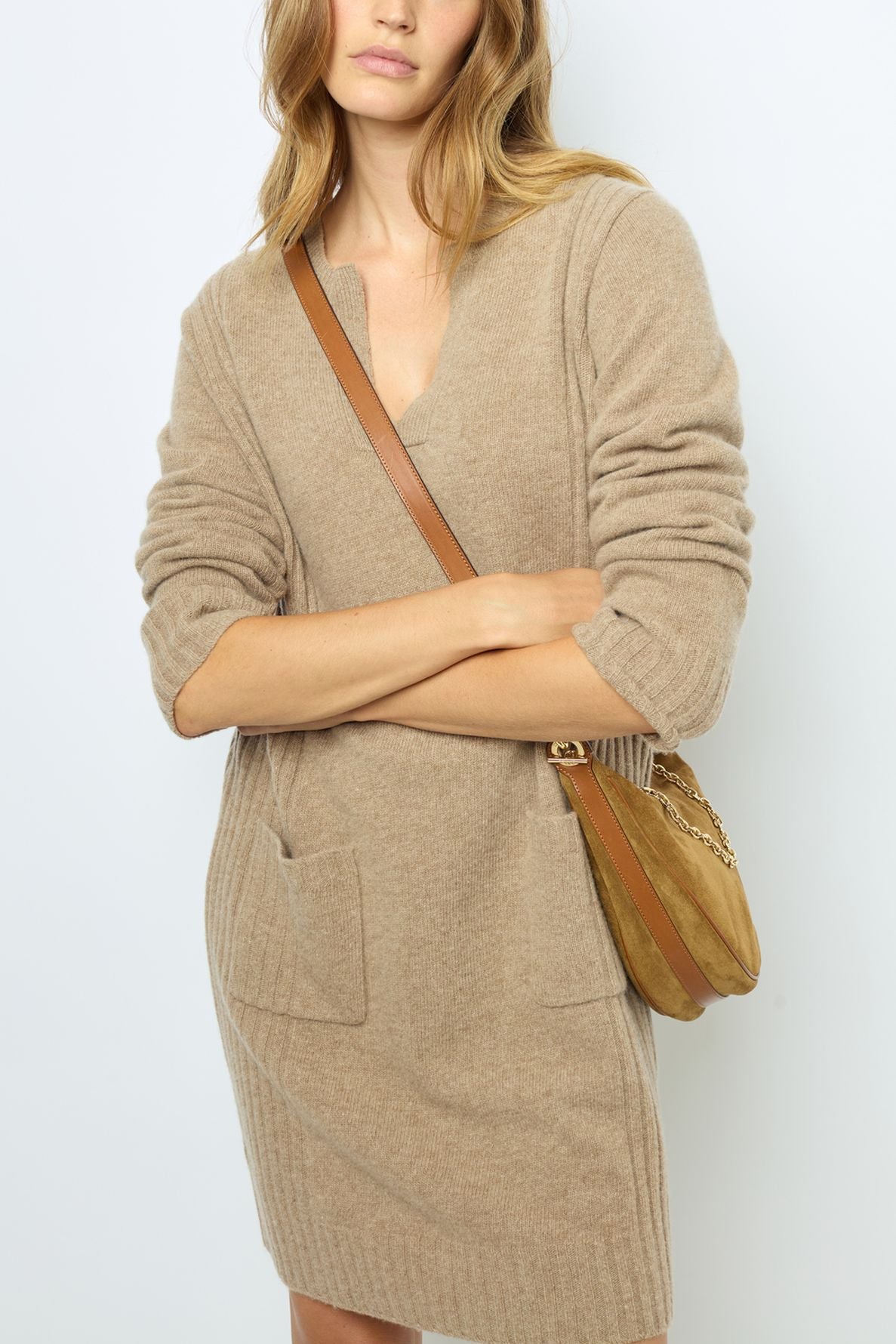 Woman wearing a beige knit dress with front pockets and a tan shoulder bag, standing against a plain light background. Her arms are crossed and her hair is wavy and loose.