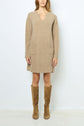 A woman wearing a tan, long-sleeve sweater dress with front pockets and knee-high suede boots stands against a plain white background.