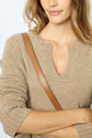 A woman with wavy light brown hair wears a beige knit sweater with a notched neckline and a brown leather shoulder strap, standing with her arms crossed.