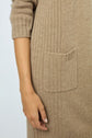 A persons hand resting by their side, wearing a ribbed, light brown knit sweater or dress with a front pocket.