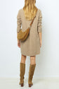 A woman stands facing away, wearing a beige sweater dress, tan knee-high suede boots, and a brown crossbody bag. Her long, wavy hair is down. The background is plain and light-colored.