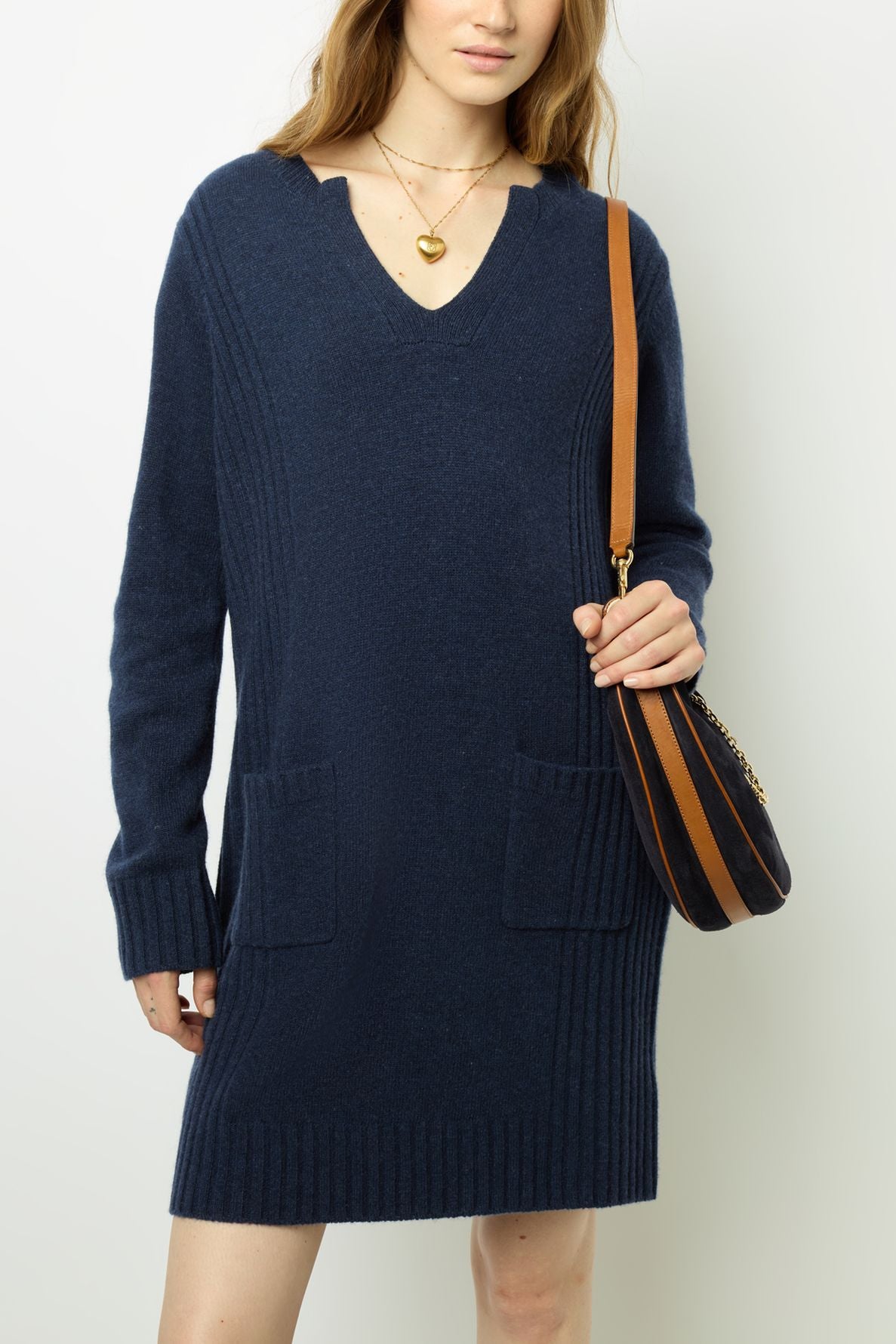 A woman wearing a navy blue robe laine sweater dress with front pockets and a gold heart necklace, holding a brown and navy striped shoulder bag, stands against a plain white background. The robe courte features a flattering col tunisien that adds a touch of elegance to her look.