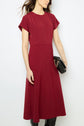 A woman wearing a short-sleeved, robe midi cintrée en microfibre rouge holds a black clutch with a chain strap. She has loose, wavy brown hair and exudes effortless silhouette glamour as she stands against a plain white background.