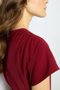 A woman with wavy brown hair wears a short-sleeved, gathered-shoulder burgundy top in soft microfibre rouge. The image is cropped to show her from the shoulders up, facing slightly away with her face partially visible, capturing an effortless silhouette glamour.