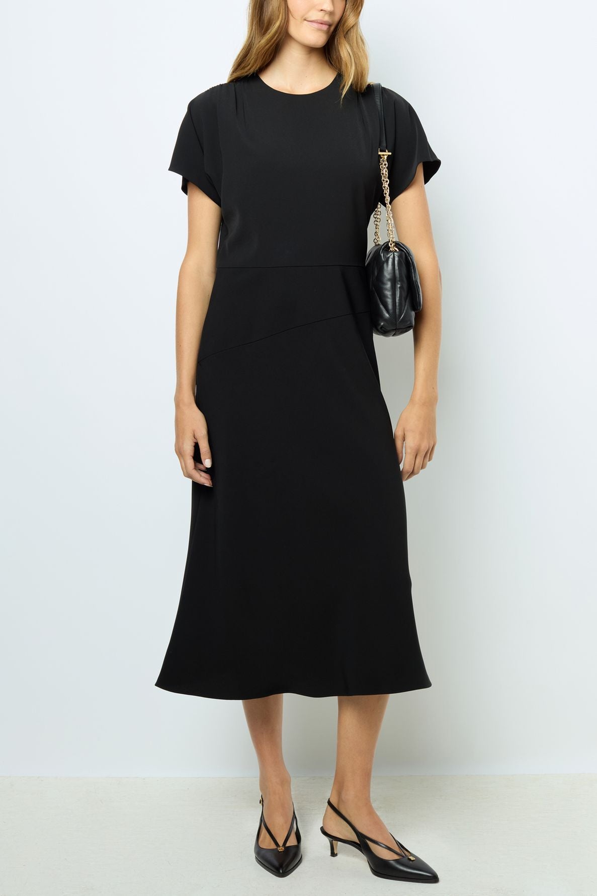 A woman wearing a robe midi élégante à manches courtes in black, paired with black pointed-toe slingback heels, and carrying a black quilted shoulder bag with a gold chain. She stands against a plain light-colored wall.