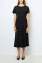 A person stands against a plain white wall, wearing a simple black robe noire cintrée, short-sleeve and midi-length, paired with black pointed-toe shoes. Their hands rest naturally by their sides.