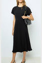 A woman wearing a robe midi élégante noire cintrée, carrying a black quilted shoulder bag, and wearing black slingback shoes stands against a plain white background. Her hair is down and she has a neutral expression.