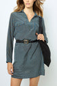 A woman wearing a blue patterned robe droite femme with a black belt and carrying a black shoulder bag. The dress features an eye-catching motif géométrique, adding a modern touch to her look. She has wavy hair and is standing against a plain light background, with her face partially out of frame.