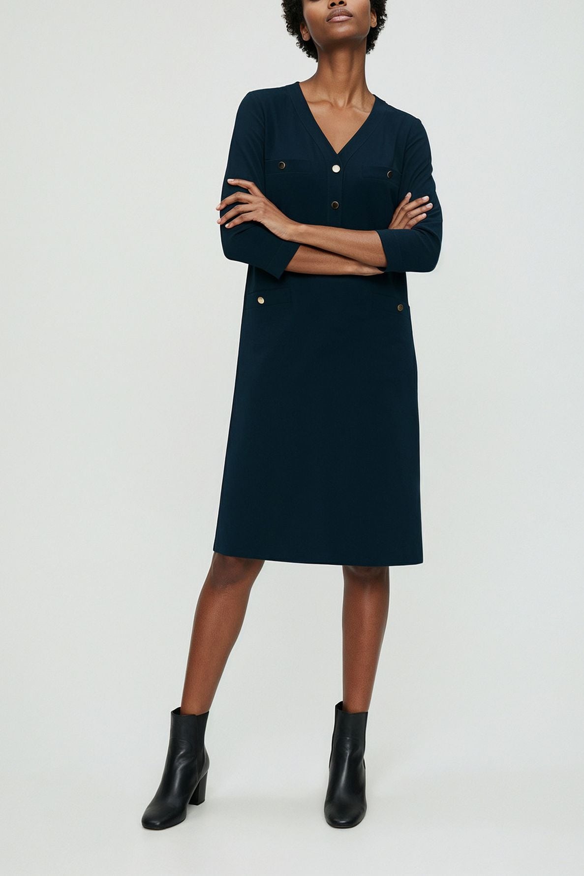 A person wearing a knee-length, navy blue robe courte with three-quarter sleeves and boutons dorés, paired with black ankle boots, stands against a plain light background.