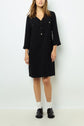 Woman wearing a chic robe courte noire adorned with boutons dorés, featuring three-quarter sleeves, white socks, and black loafers. She stands against a plain white background. Her hair is straight and loose, and her face is partially visible.