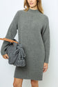 Woman wearing a long-sleeved gray knit sweater dress with col montant holds a large gray knit bag with tassels and a tan leather strap. Background is plain white.