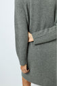 A person wearing a long, gray, ribbed sweater with un col montant stands with one arm bent behind their back, holding their opposite elbow. The image is cropped to show only the torso and upper legs.