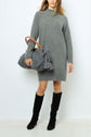 Woman in a gray knit sweater dress with col montant and black knee-high boots holds a large gray knit bag with wooden handles and tassels, standing against a plain white background.
