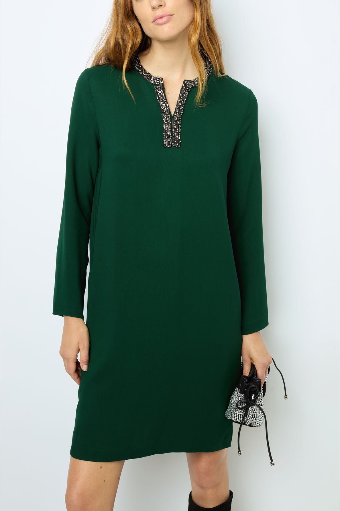 A woman wears a vert profond long-sleeved dress with a col strass and holds a small black and white purse.