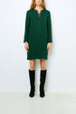 A person stands against a white background wearing a robe courte vert profond with a patterned neckline, featuring a chic col strass, paired with black knee-high boots.