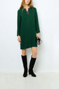 A person wearing a robe courte vert profond with a col strass, paired with black knee-high boots and holding a small black and silver purse, standing against a plain white background.