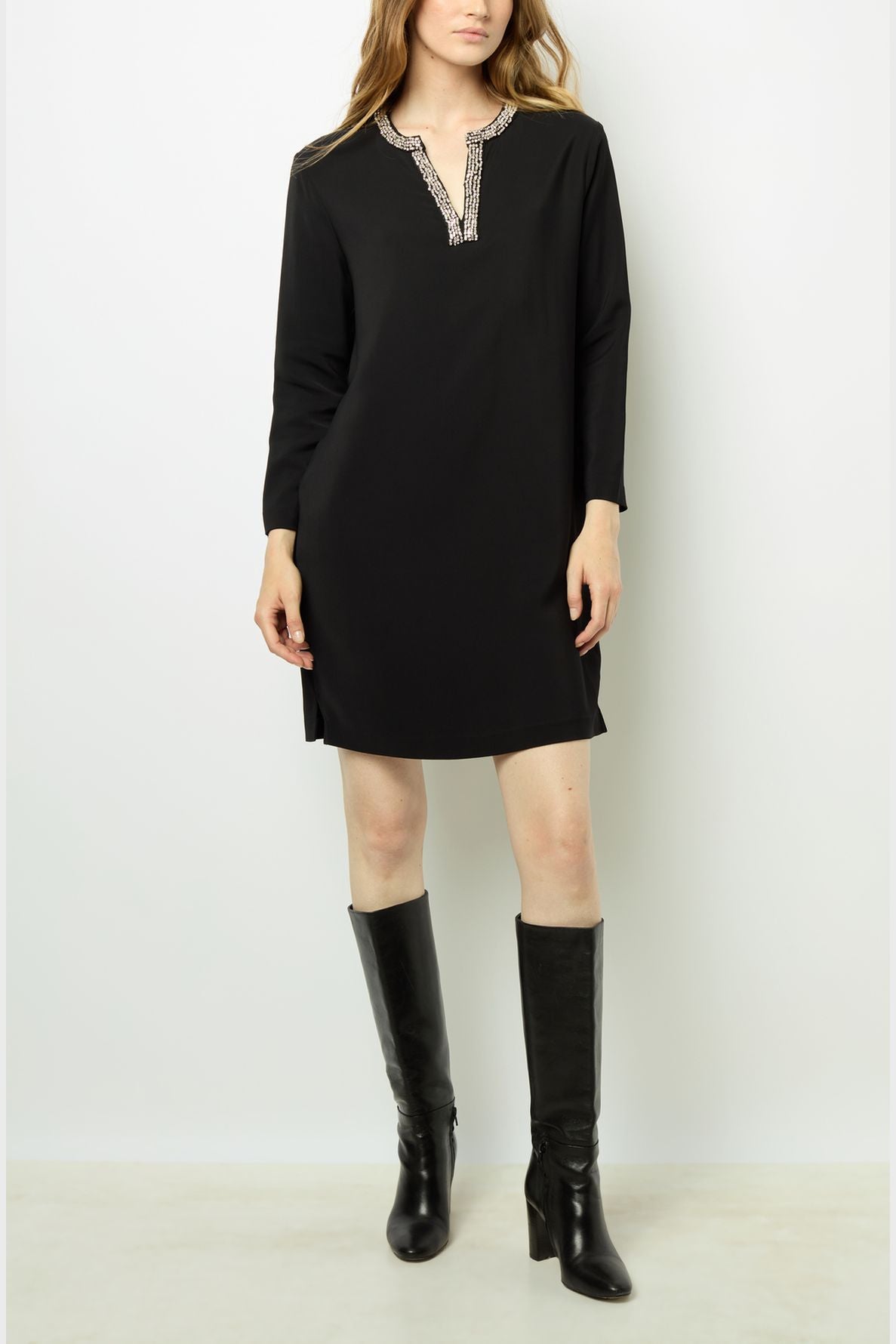 A person wearing a robe courte noire à manches longues with a col strass and knee-high black boots stands against a plain white background.