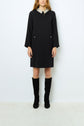 Woman wearing a black, long-sleeve robe courte with a strass-embellished col chemise and front pockets, paired with black knee-high boots, standing against a plain light background.