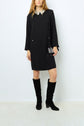 Woman wearing a black robe courte with a col chemise adorned with strass, paired with black knee-high boots and holding a black crossbody bag with silver accents, standing against a plain white background.