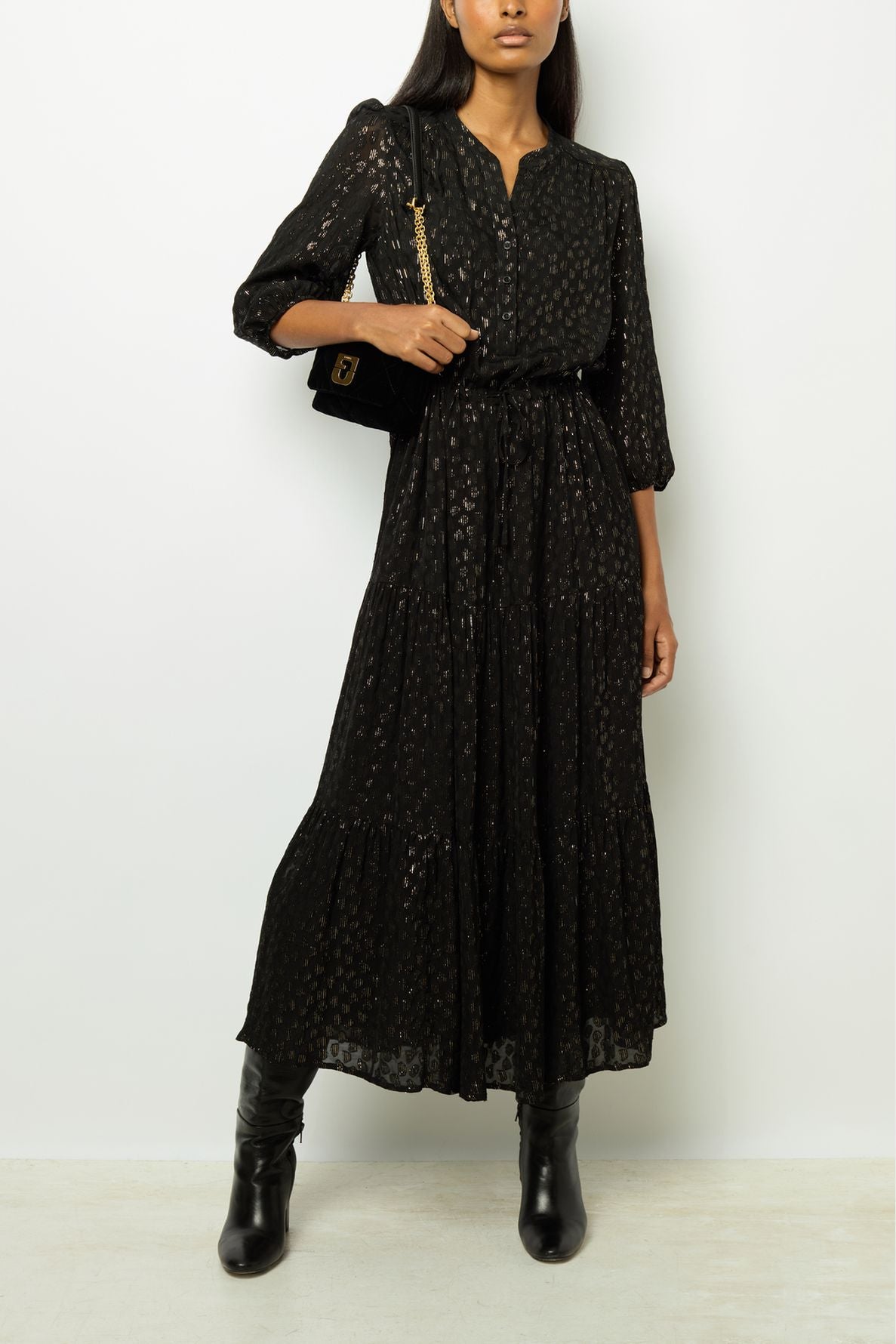 Woman wearing a robe longue noire with a subtle lurex shimmer and three-quarter sleeves, its taille ajustable creating an elegant silhouette, paired with black knee-high boots and a black shoulder bag with a gold chain strap, standing against a plain white background.