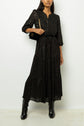 Woman wearing a robe longue noire with a subtle lurex shimmer and three-quarter sleeves, its taille ajustable creating an elegant silhouette, paired with black knee-high boots and a black shoulder bag with a gold chain strap, standing against a plain white background.