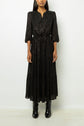 A woman wearing a robe longue noire with a subtle lurex pattern and three-quarter sleeves, paired with black ankle boots, stands against a plain white background.