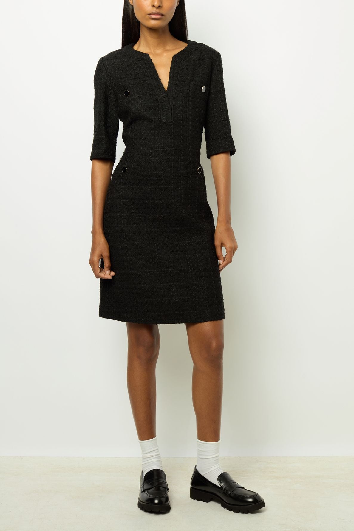 A person wearing a knee-length black textured dress with short sleeves, white socks, and black loafers stands against a plain white background. Their arms are relaxed at their sides.