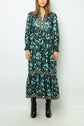 Woman wearing a long-sleeved, teal and black floral jacquard maxi dress with a V-neck and tiered hem, paired with black boots, standing against a plain white background.