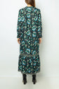 Woman wearing a long-sleeved, ankle-length robe midi with a teal, black, and white abstract floral imprimé végétal pattern, standing with her back to the camera; black heeled boots are visible.