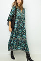 Woman wearing a long, patterned teal and black robe midi with an imprimé végétal design and a deep V-neck, paired with black heeled boots and a large black shoulder bag, standing against a plain white background.