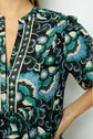 A person wearing a dark jacquard blouse with green, blue, and white floral designs stands with one hand on their hip, showing the upper torso and part of the arm.