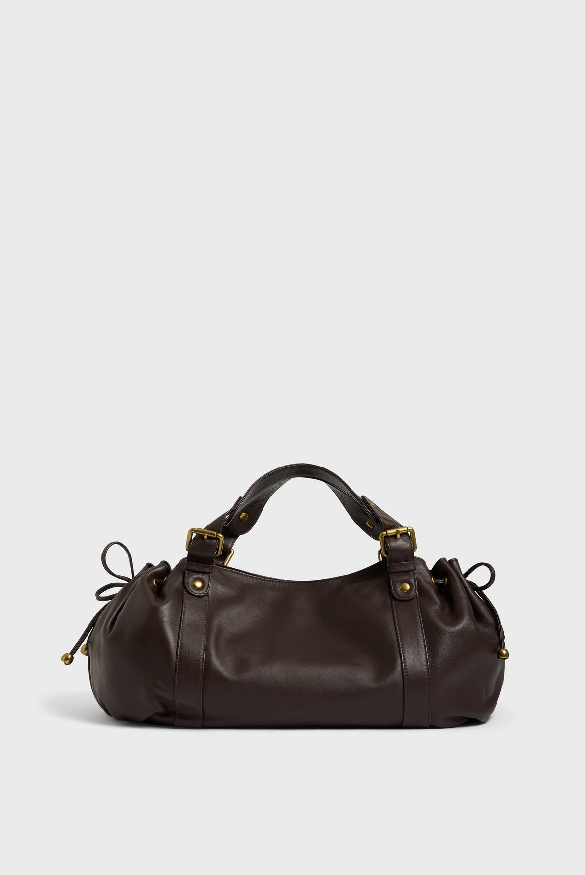 A dark brown cuir délicat leather sac à main with gold hardware, adjustable straps, and side drawstrings, photographed on a plain light gray background—a refined piece of maroquinerie Darel craftsmanship.