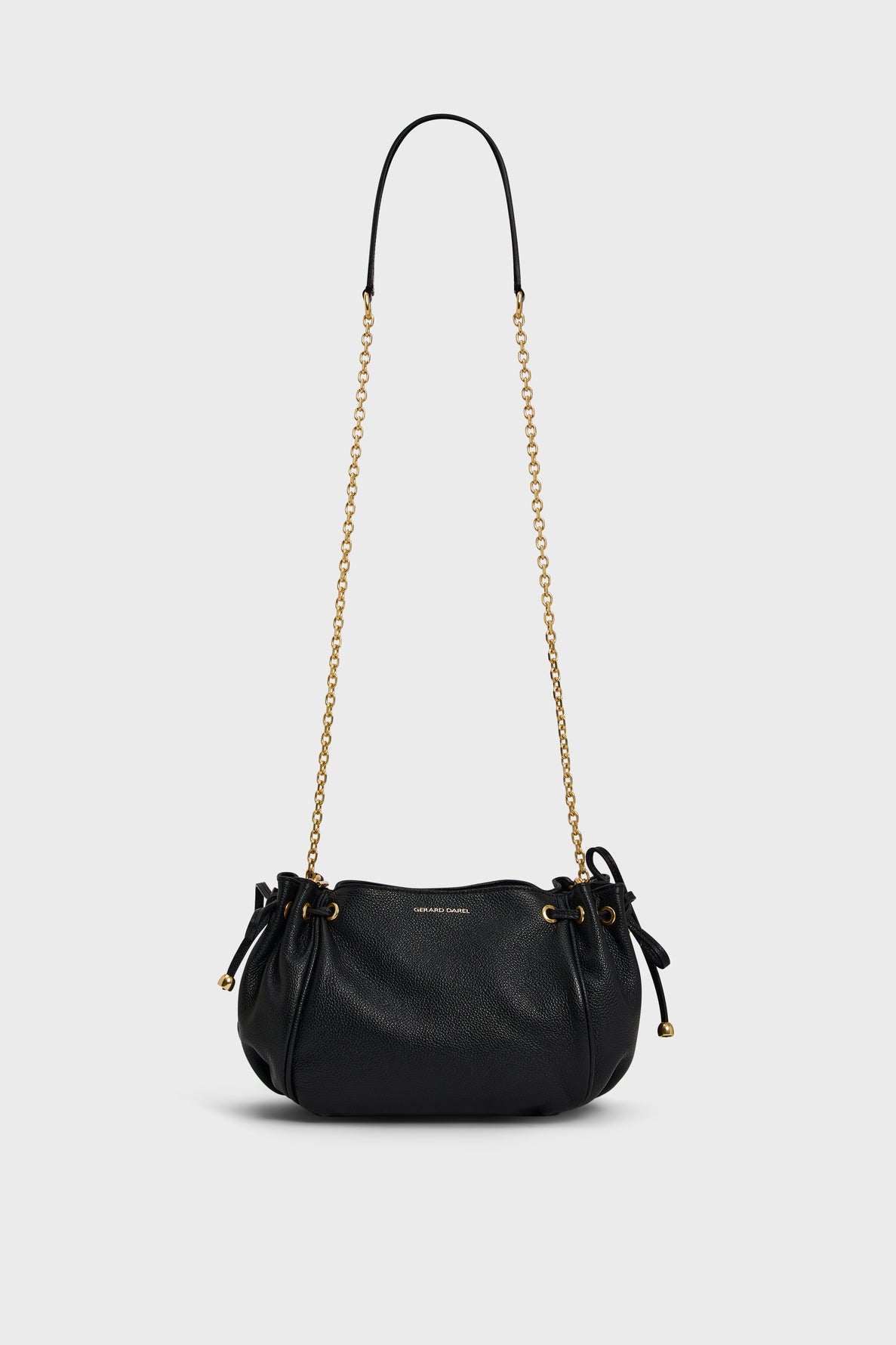 A small black cuir grainé leather sac bandoulière with a gold chain strap, drawstring sides, and subtle gold Gerard Darel branding on the front, shown on a plain light background.