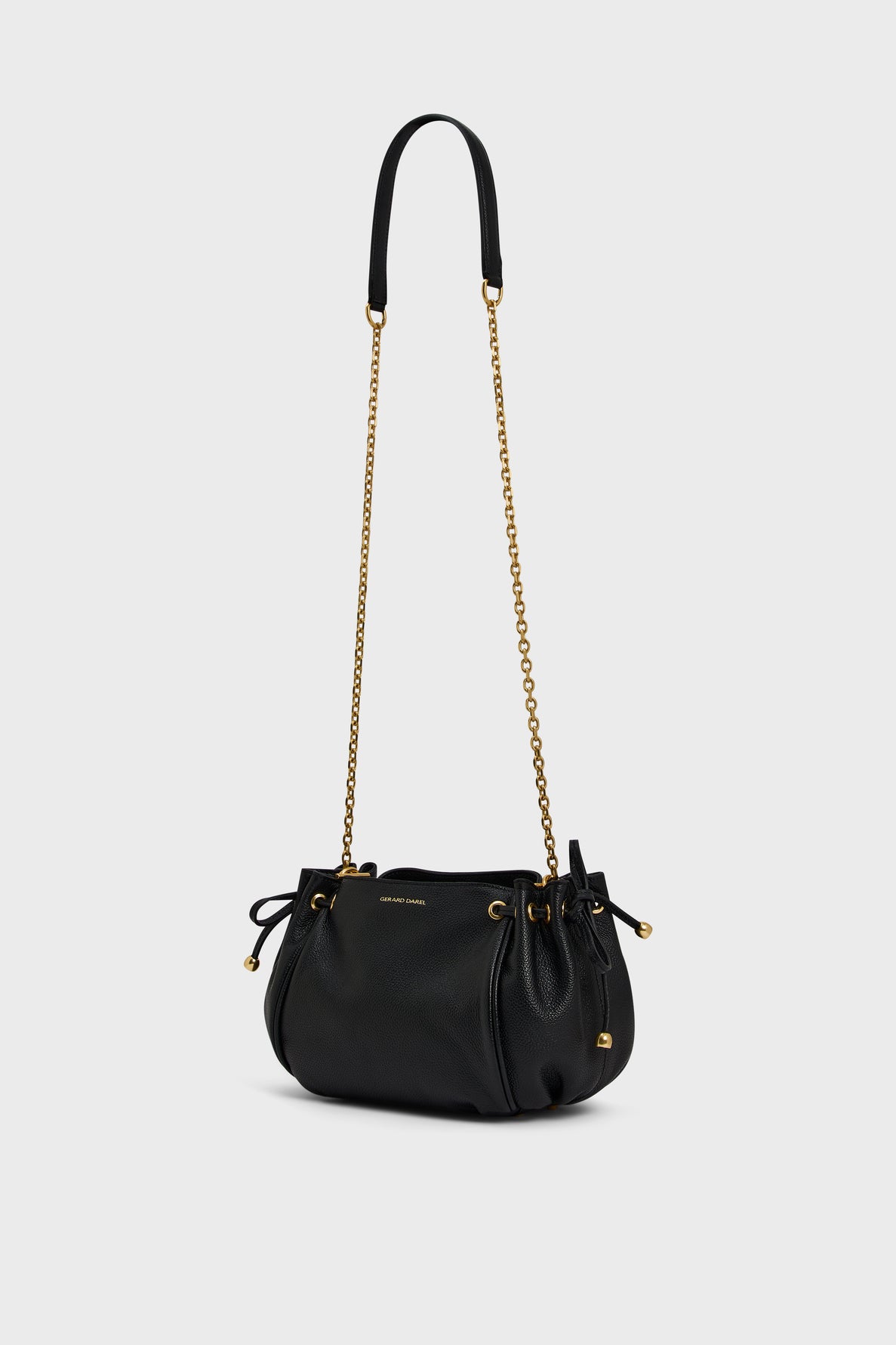 Black leather drawstring sac bandoulière by Gerard Darel, featuring gold chain and shoulder strap, gold hardware, and subtle branding displayed on the front. Crafted from luxurious cuir grainé, the bag has a soft, rounded shape and is set against a plain light background.