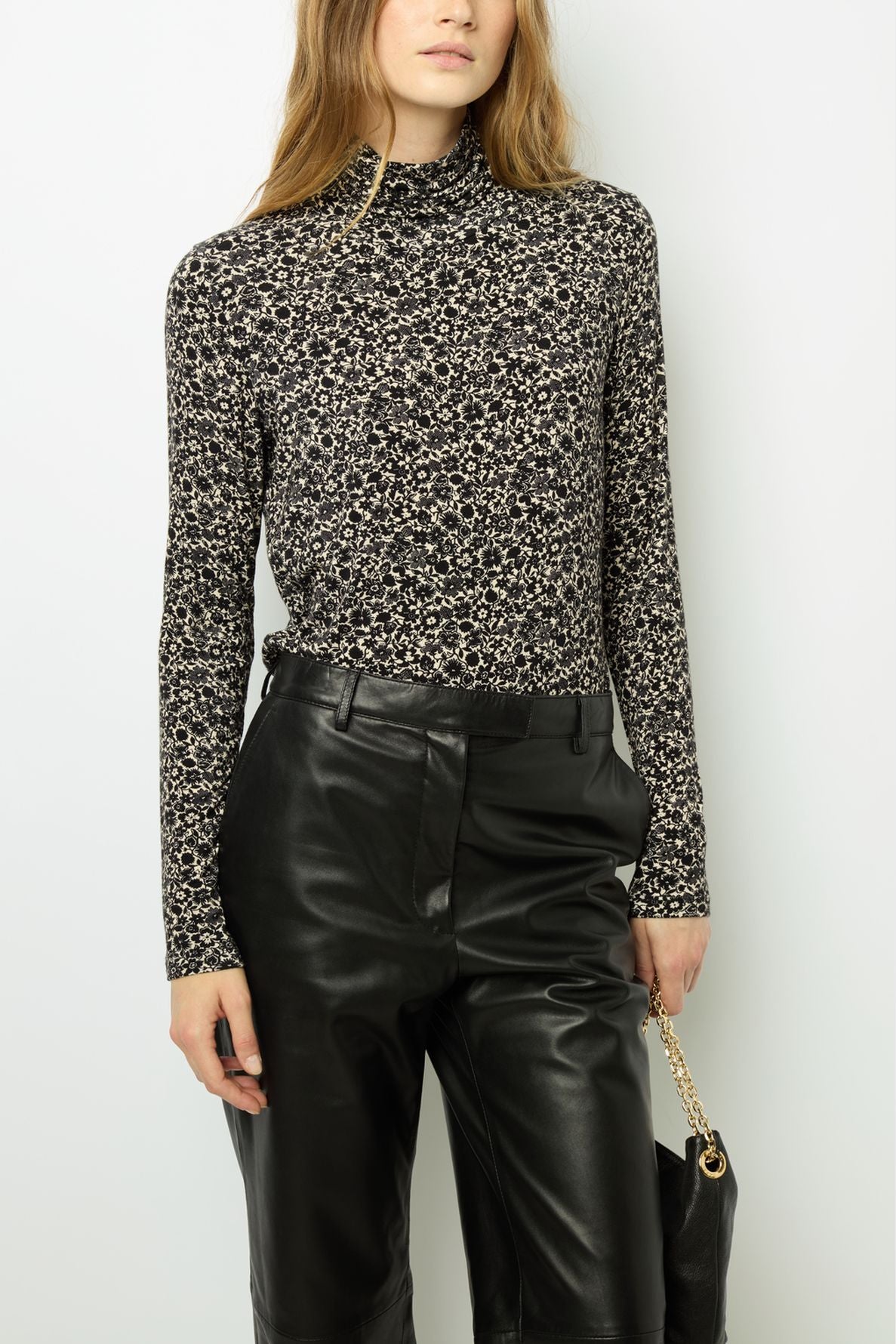 A person wearing a black and white floral long-sleeve mock-neck top with high-waisted black leather pants, holding a black handbag with a gold chain strap.