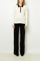 Person wearing a long-sleeve polo blanc en coton with a sleek col noir and buttons, paired with black wide-leg pants featuring a subtle circular pattern and black shoes. The background is plain white.